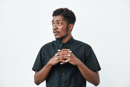 Young black man with dark skin wearing black shirt standing with hands clasped looking sideways on white background. Portrait of thoughtful African American male in casual clothing.の写真素材