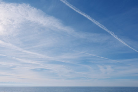 Clear blue sky over a calm ocean, intersecting wispy clouds and contrails, expansive horizon, serene...