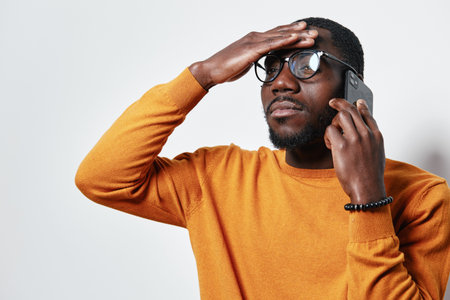 African American man with dark skin wearing glasses and orange sweater talking on smartphone with...