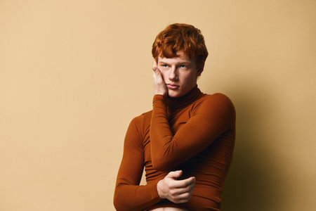 Young man with red hair and freckled skin wearing brown turtleneck posing thoughtfully against beige...