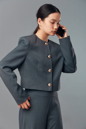Woman in gray suit standing and talking on mobile phone with serious expression in studio. Female...