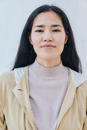Calm woman with straight dark hair wearing a beige jacket over a light sweater against a pale wall,...