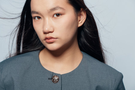 Young woman portrait with long black hair and serious expression wearing gray jacket in studio....