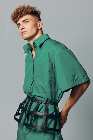 Young man with short curly hair in green oversized shirt and wide belt posing against gray background in studio. Male fashion model shows modern streetwear style and casual outfit profile.の写真素材