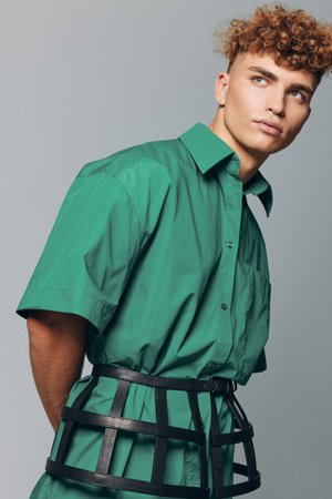 Young man with curly hair wearing green shirt and black cage belt posing against gray background in studio for fashion portrait and style concept.の写真素材