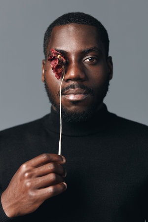 Creative conceptual portrait of African man holding dried flower in front of eye with serious expression, minimalistic visual style and dark clothing on gray background.の写真素材