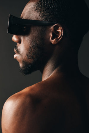African man with dark skin wearing futuristic sunglasses in creative conceptual visual style, closeup portrait with bare shoulders and textured skin on dark background.の写真素材