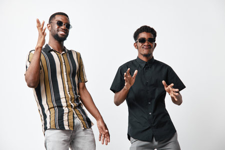Two young African American men with dark skin wearing sunglasses and casual shirts posing and smiling on white background in studio for lifestyle and friendship.の写真素材