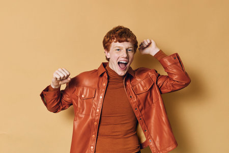 Young man with red hair wearing brown leather jacket and sweater, smiling and raising fists in excitement against beige background. Casual style and energetic mood.の写真素材