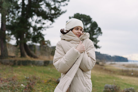 Woman in a beige quilted coat and knit beanie stands in an open outdoor landscape, wrapped in a...
