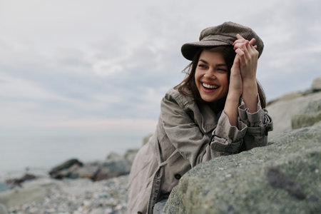 Smiling young woman in casual clothes and hat leans on rock near rocky beach under cloudy sky....