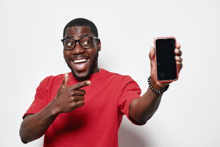African American man with dark skin wearing glasses and red shirt smiling and pointing at smartphone screen for technology and communication concept on white background.の写真素材