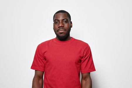 African American man with dark skin wearing red t shirt standing against white background looking at...