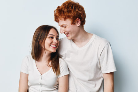 Smiling young couple with red hair and brunette looking at each other with happy expressions in casual white t-shirts on light background studio.の写真素材