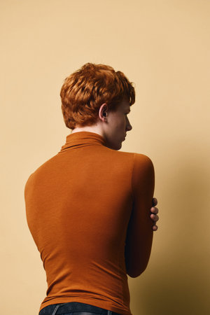 Young man with red hair wearing orange long sleeve sweater standing back to camera posing against...