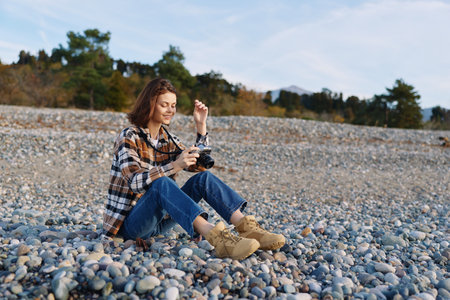 Woman sitting on a pebble beach in casual outdoor attire, focused on a camera, surrounded by...