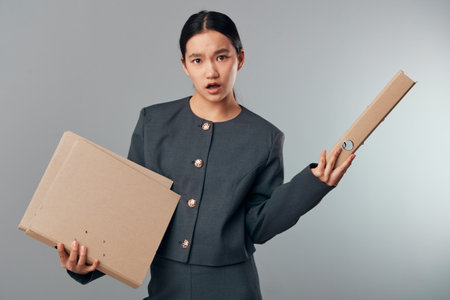Woman holding cardboard boxes with confused expression wearing formal jacket. Delivery, packaging, logistics and shipping concept with uncertain female in gray outfit on plain background.の写真素材
