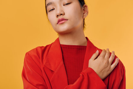 Young woman with closed eyes wearing red jacket and sweater posing with hand on shoulder against yellow background in studio portrait for fashion and emotion.の写真素材