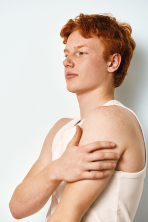 Young man with red hair and pale skin wearing white tank top touching shoulder in studio. Side view...