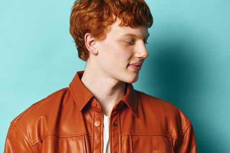 Young man with red hair and freckles wearing a brown leather jacket looks sideways against a blue background in a studio portrait, casual style and thoughtful expression.の写真素材