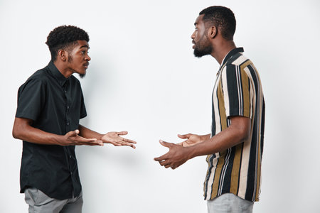 Two black men with dark skin having a serious conversation in casual clothes on white background. African ethnicity, communication, discussion, disagreement, standing indoors.の写真素材