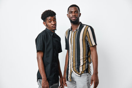 Two young black men with dark skin standing together in casual shirts against white background. African ethnicity, friendship, confidence, and modern style in studio portrait.の写真素材