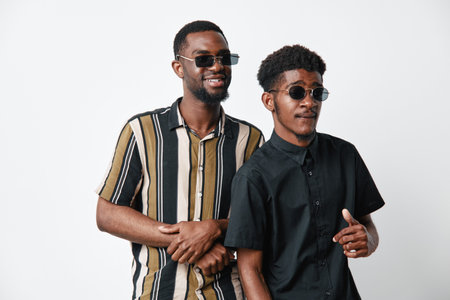 Two African men with dark skin wearing sunglasses and casual shirts standing together on white...