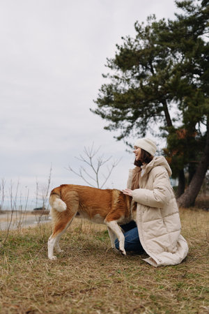 Woman kneeling on a grassy meadow outdoors, petting a dog in a warm winter coat, with trees and a...