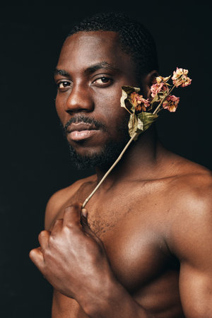 Black man with dark skin holding dried flowers near face in creative conceptual visual style portrait with serious expression on dark background.の写真素材