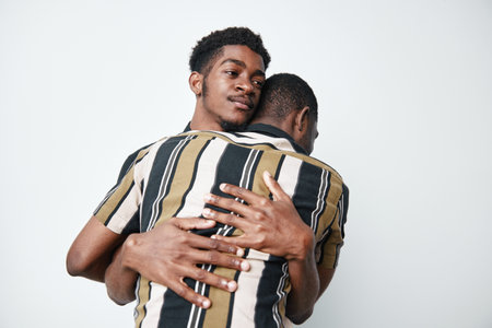 Two young black men with dark skin hugging each other in emotional embrace on white background. Male friends showing support, connection, and affection with casual striped shirts.の写真素材