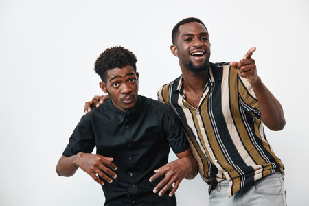 Two young black men with dark skin posing together on white background, casual clothing, smiling and pointing, friendship and happiness concept.の写真素材