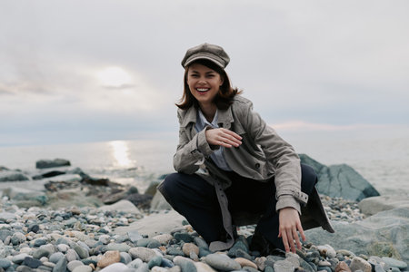 Smiling young woman in casual clothes and hat crouches on rocky beach near calm sea. Overcast sky and soft natural light create peaceful mood for outdoor lifestyle and travel concept.の写真素材