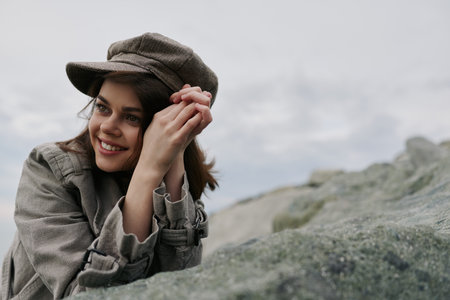 Smiling young woman wearing a patterned coat and matching cap leans on a rocky surface outdoors. The cloudy sky background creates a natural and relaxed mood in the image.の写真素材