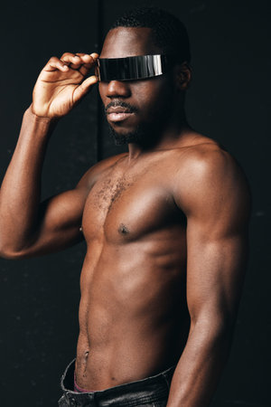African man with muscular body and dark skin wearing futuristic visor glasses in creative conceptual visual style. Portrait of confident male model with bare chest and modern accessory.の写真素材