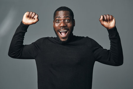 African man with dark skin wearing black sweater raising fists in excitement and joy in creative conceptual visual style isolated on gray background.の写真素材