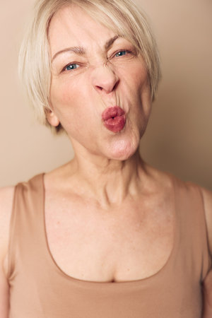 Close-up of an elderly woman making a playful face with puckered lips and squinted eyes. Natural skin texture and short blond hair visible against neutral background.の写真素材
