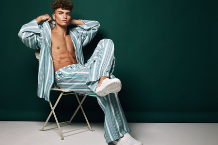 Young man sitting on chair with striped pants and open shirt in studio with dark green background. Casual fashion pose with confident expression and relaxed posture.の写真素材