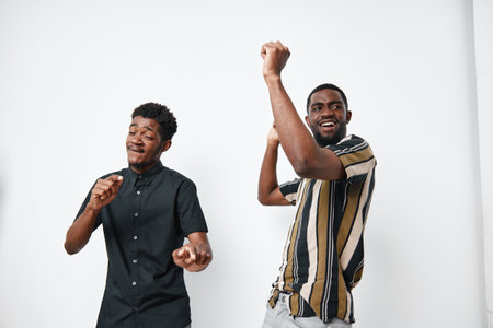 Two African men with dark skin dancing and enjoying music together in casual clothes on white background. Friends having fun and expressing happiness indoors.の写真素材