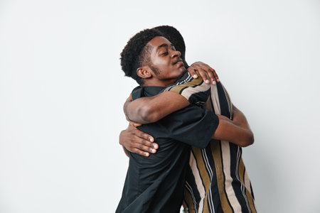 Two men with dark skin hugging each other in a friendly embrace on white background. African ethnicity, casual clothing, emotional connection, support and friendship concept.の写真素材