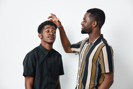 Two African American men with dark skin interacting in studio, one playfully touching the others hair, casual clothing and friendly expression on white background.の写真素材