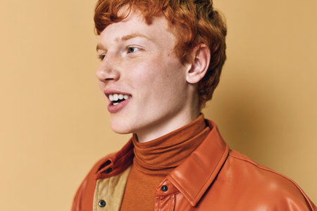 Young man with red hair wearing orange turtleneck and leather jacket smiling looking aside on beige background in studio portrait with natural emotion and casual style.の写真素材
