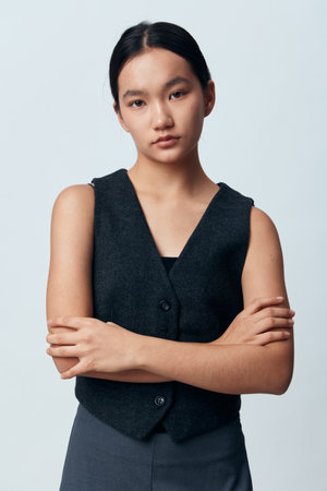 Young woman with straight black hair wearing dark vest and pants standing with arms crossed on light background in studio. Serious expression, isolated, casual fashion and confident pose.の写真素材