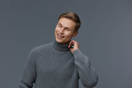 Smiling happy young caucasian man wear gray turtleneck sweater look aside positive emotion isolated on dark gray background cozy winter style mental health and gadgets conceptの写真素材