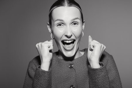 Excited Caucasian woman with light skin expresses joy with clenched fists and open mouth on grey background studio portrait.の写真素材