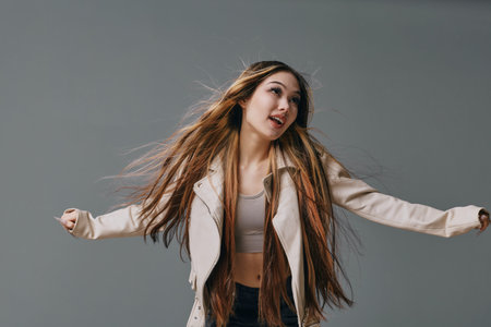 Young woman with long hair is dancing in casual beige jacket and crop top against gray background. Movement, joy and fashion style in studio for lifestyle and youth culture.の写真素材