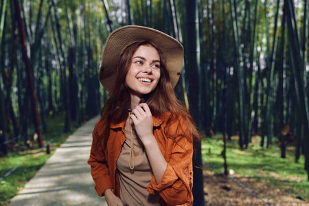 Woman with a warm smile wearing a straw hat walks along a forest path, portrait-style photo of a young brunette in casual jacket, sunlight and relaxed outdoor nature lifestyle.の写真素材