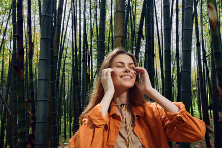 Woman in a bamboo forest smiling with eyes closed, enjoying nature and relaxation. Casual model in orange jacket breathes fresh air among tall green trees in a serene grove.の写真素材