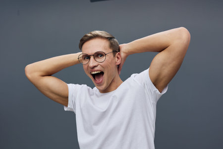 Excited young caucasian man wearing glasses and white t-shirt looking camera isolated on gray background expressing joy and surprise digital nomad conceptの写真素材