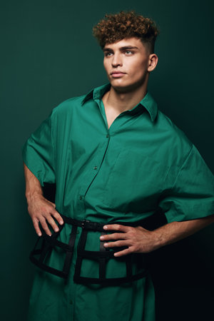 Young man with curly hair wearing oversized green shirt and black belt posing with hands on hips against dark green background in studio portrait.の写真素材