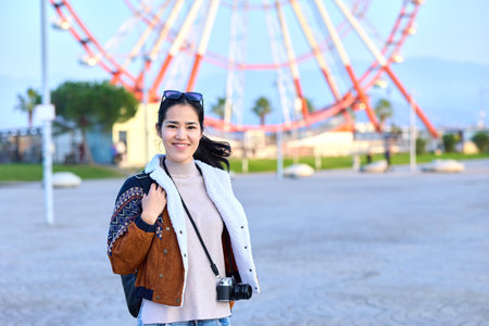 Woman with camera in outdoor amusement park, smiling and casual, carrying a bag, ferris wheel in the background, sunny day, travel vibe, candid street photography, lively and natural sceneの写真素材
